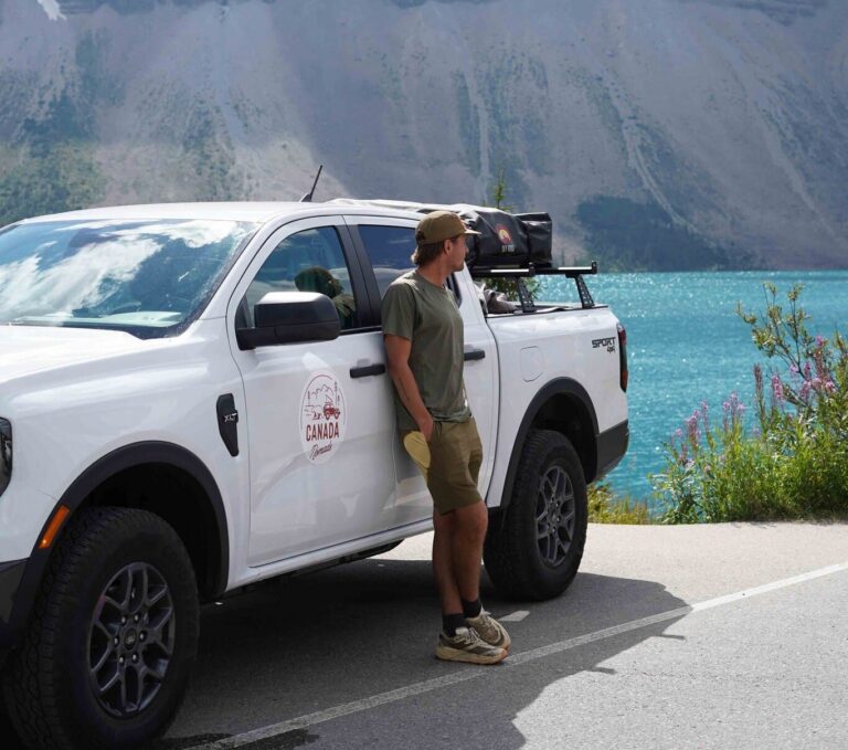 man leaning on white truck by turquoise lake and mountains